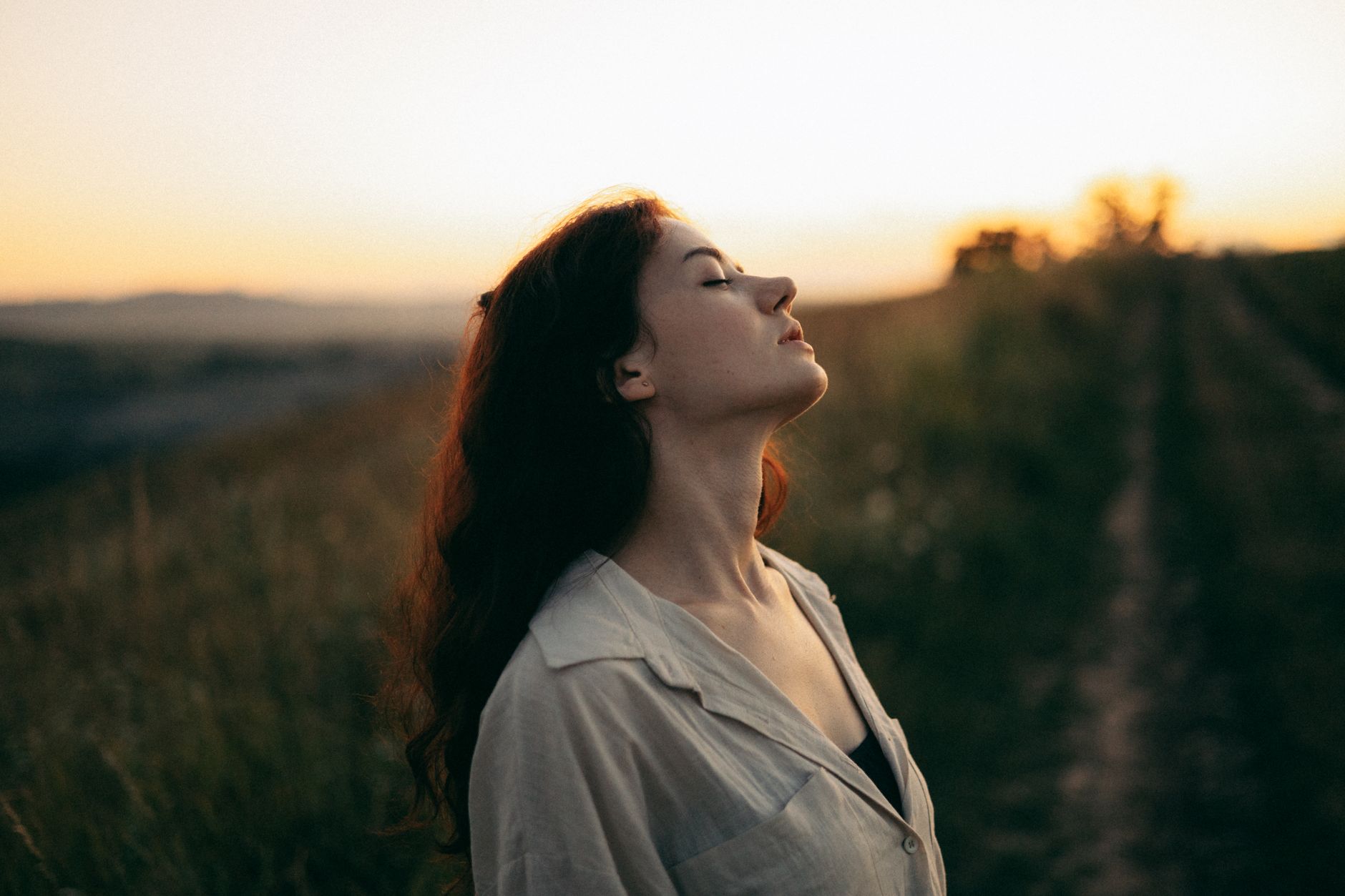 Woman standing with eyes closed in a field during sunset, exuding tranquility and peace.